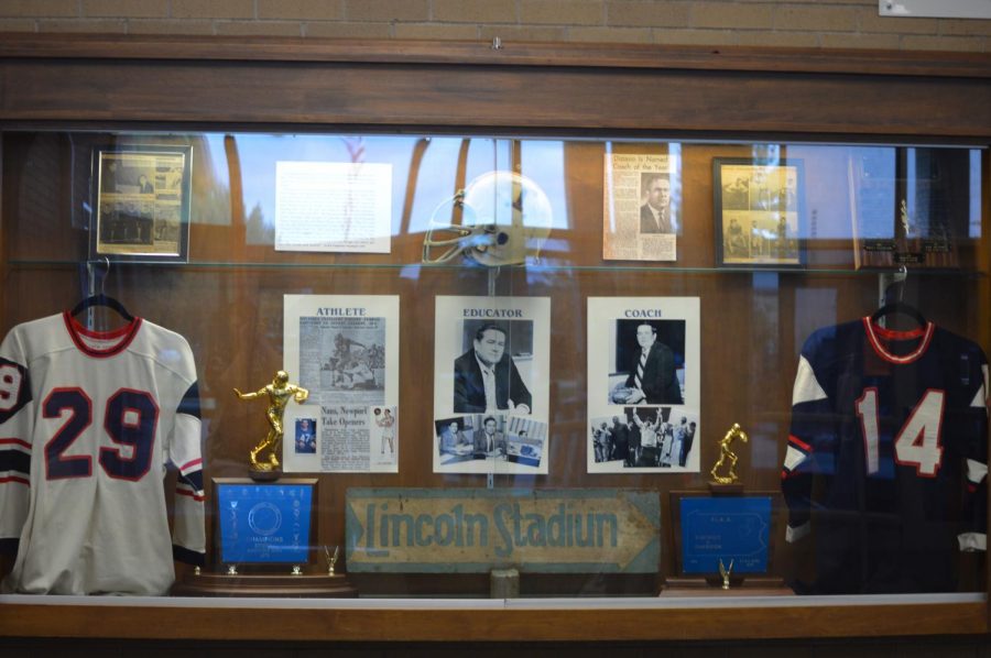 Septembers historical display case features two old Trojan team jerseys and an old helmet. It also displays pictures of Coach Distasio and two football championship trophies. Additionally, there are also the Coach Distasio Memorial Award along with old pictures of football players.